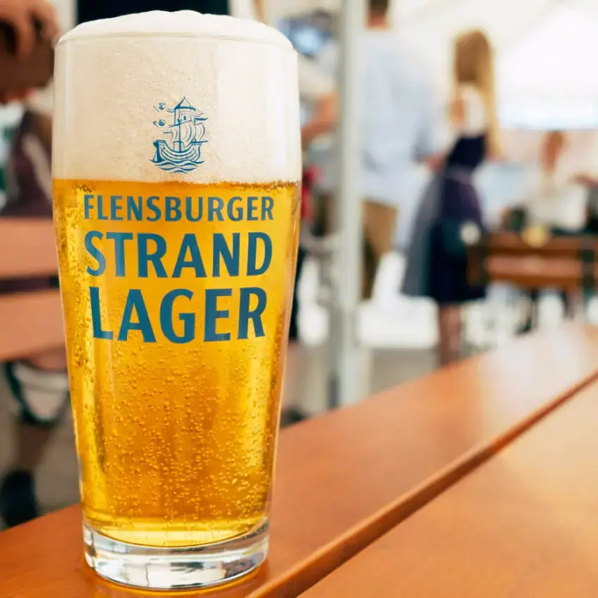 Ein Glas Flensburger-Strand-Lager-Bier auf hölzernem Biertisch im belebten Biergarten.