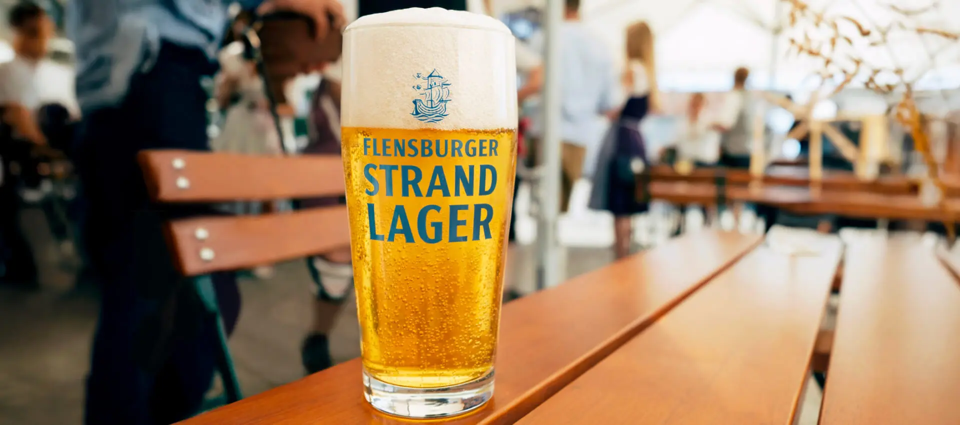 Ein Glas Flensburger-Strand-Lager-Bier auf hölzernem Biertisch im belebten Biergarten.