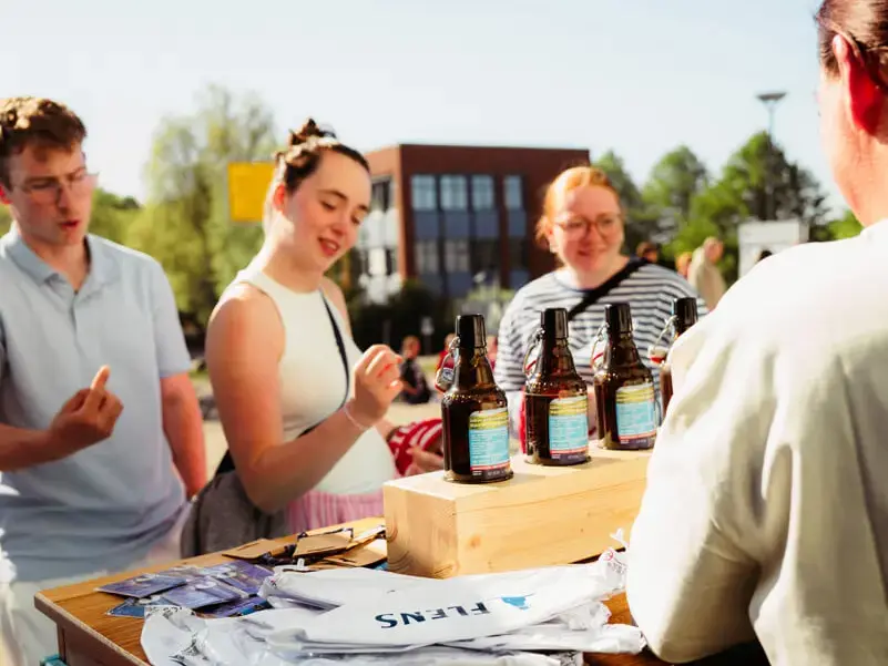 Promostand von FLENS im Freien mit Bierflaschen auf Holzblock, Promoter und Besucher bei Verkostung und Prömpelspiel.