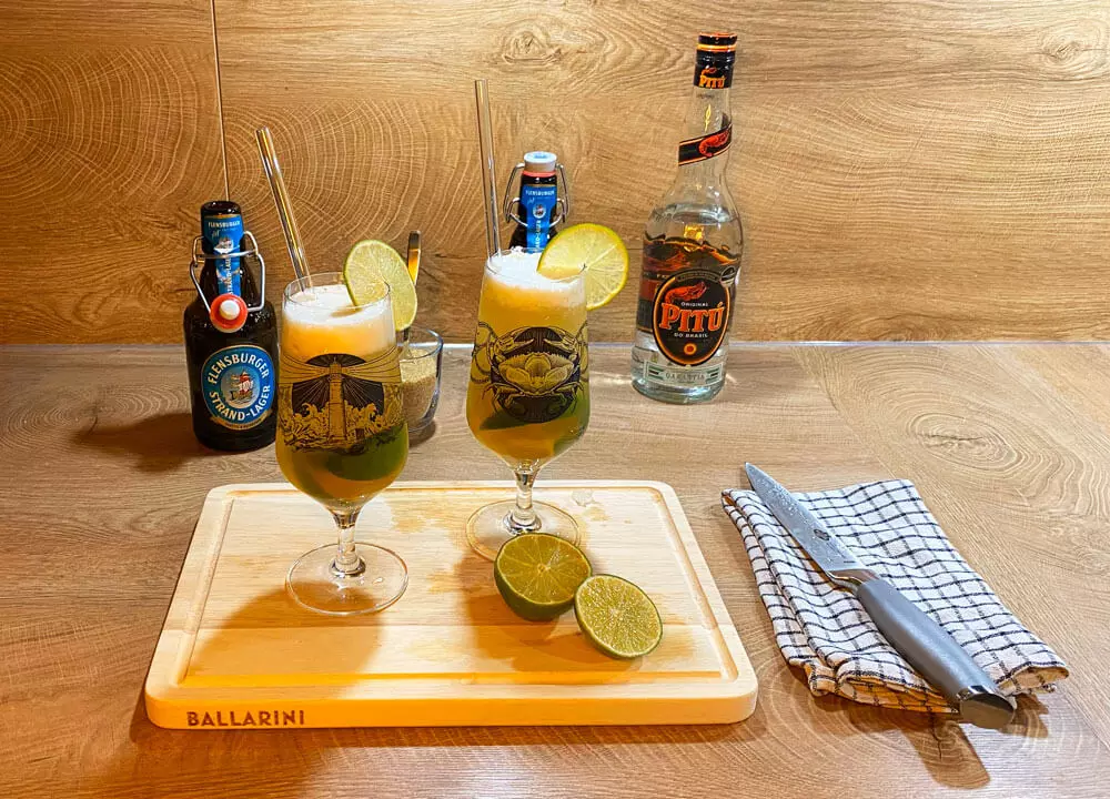 Zwei Gläser vom fertigen Biercocktail Caibierinha mit Flensburger Strand-Lager, weißem Rum und Limette, dahinter Cocktailutensilien.