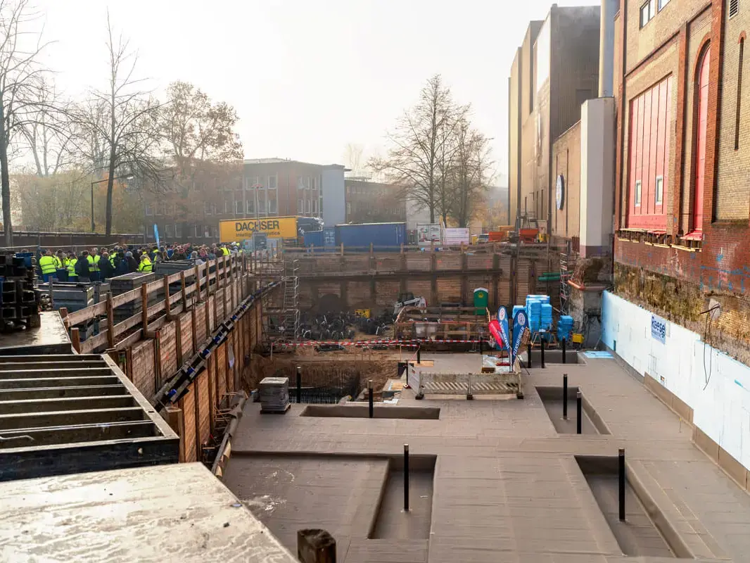 Belegschaft steht oben vor der Baustelle des neuen Sudhauses für die Flensburger Brauerei neben dem historischen Brauereigebäude bei Tageslicht.