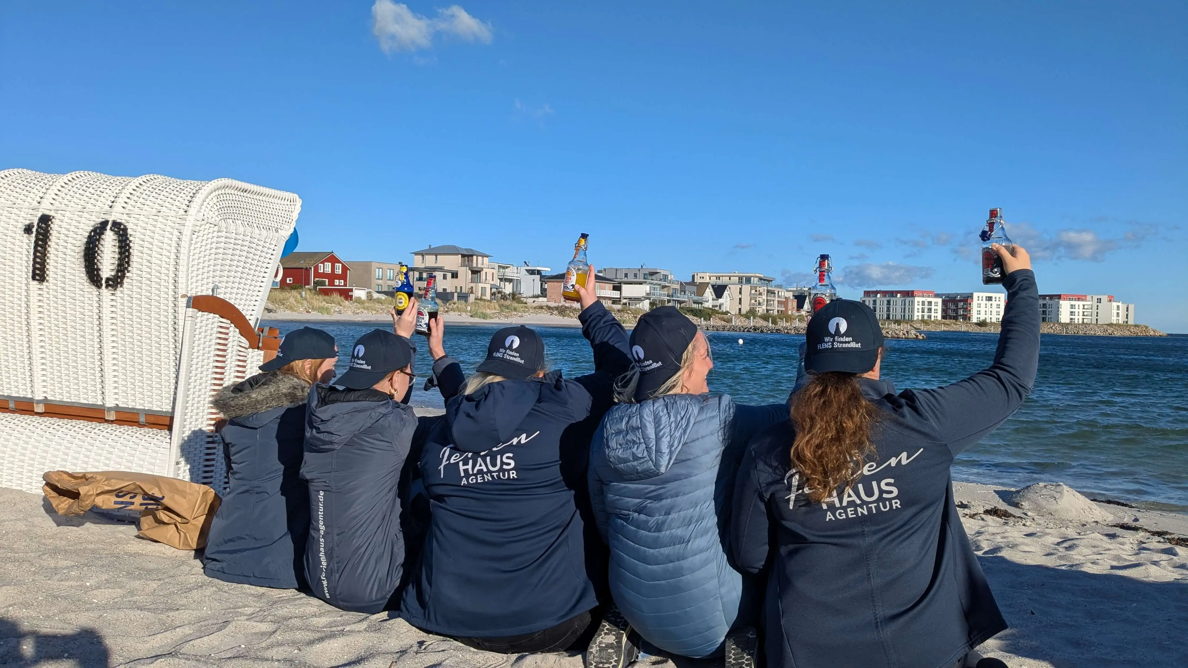 Gruppe von Freiwilligen der FLENS StrandGut Aktion, alle in blau gekleidet, sitzt am Strand hebt FLENS Flaschen für ein Foto mit Meer im Hintergrund.