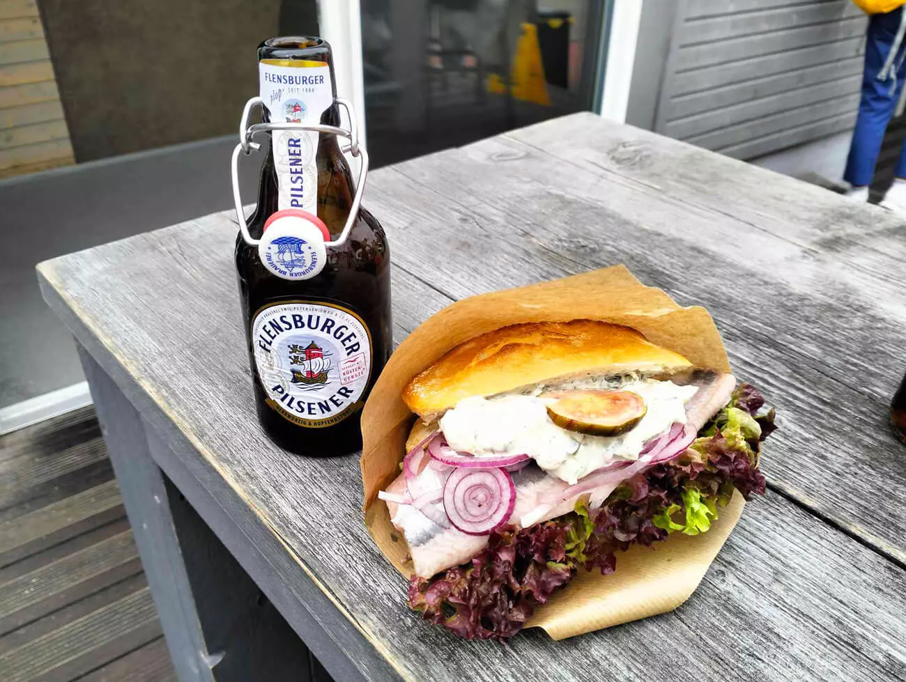 Auf einem grauen Holztisch ein Fischbrötchen und dahinter eine Flasche FLENS.