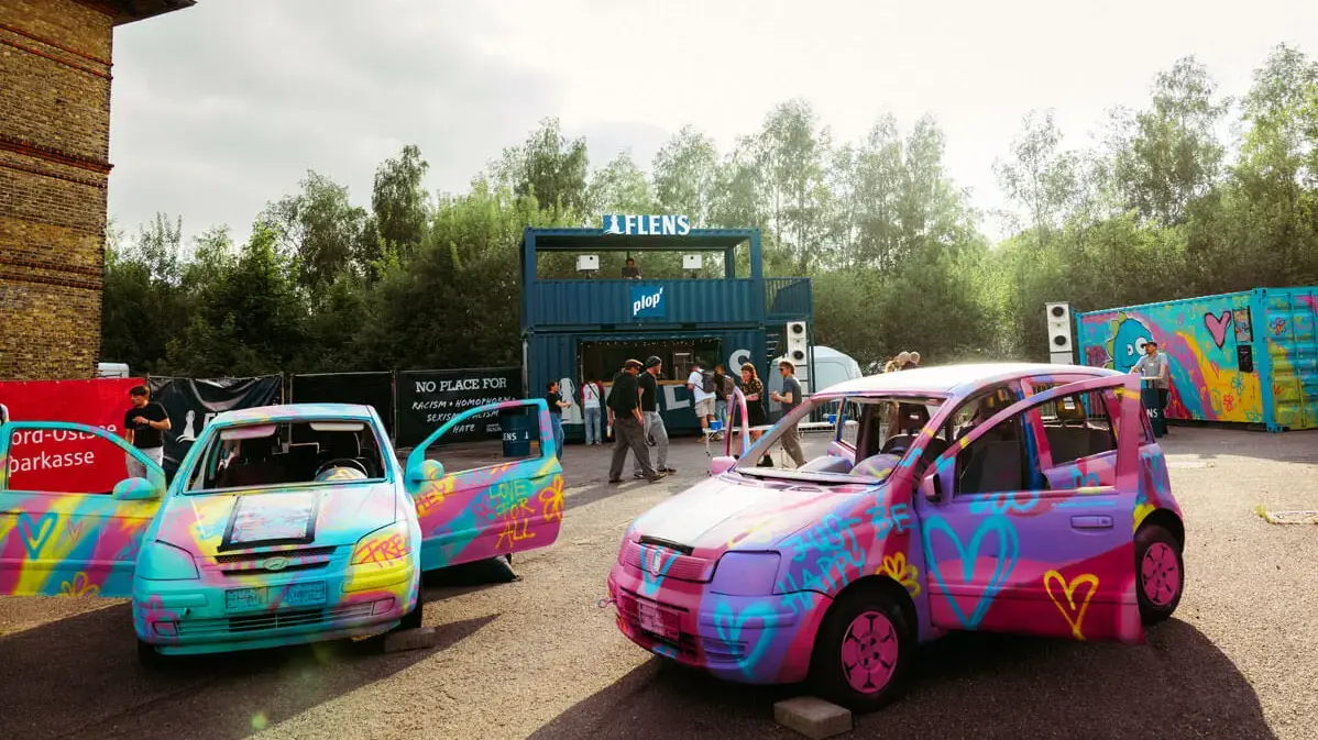 Bunte Graffiti-Autos mit Herzen auf Festivalgelände vor blauer FLENS Containerbühne, Menschen im Hintergrund.