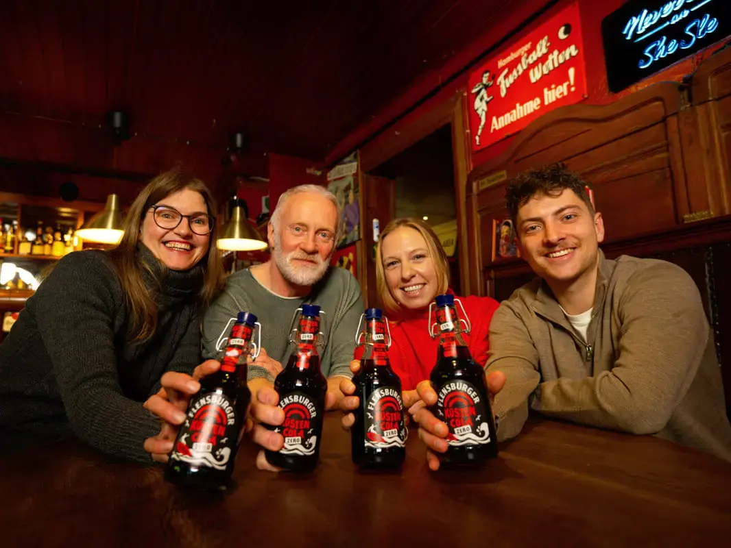 Eine Gruppe bunt gemischter Leute in einer Bar hält die Flensburger Küstencola Zero in die Kamera.