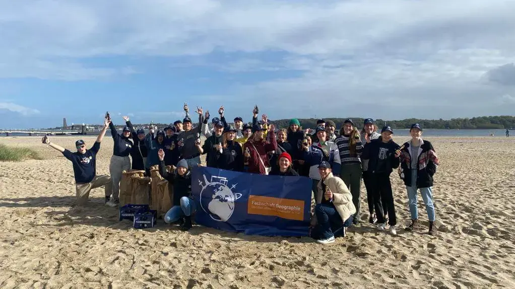 Gruppe von Freiwilligen jubelt mit Banner am Strand bei sonnigem Wetter bei einer FLENS StrandGut Aktion.