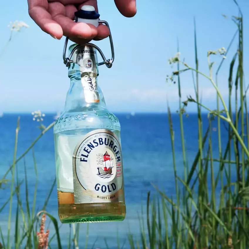 Bierflasche Flensburger Gold wird am Bügelverschluss gehalten, im Hintergrund eine grüne Wiese vorm Wasser.