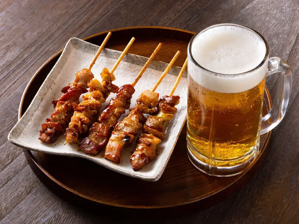 Yakitori (gegrillte Hähnchenspieße) auf Teller mit Bierglas daneben auf Holztisch.