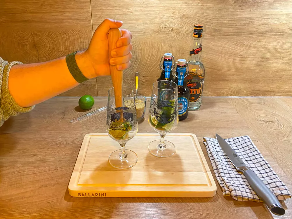 Mit einem Stößel werden die Limettenachtel im Glas zerstoßen, dahinter Cocktailutensilien.