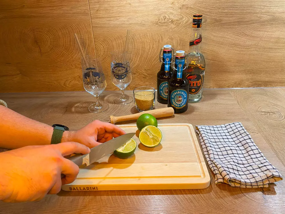 Eine Limette wird auf einem Holzbrett geschnitten, dahinter Cocktailutensilien.