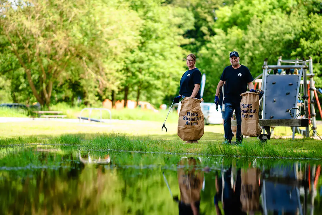 Zwei Freiwillige reinigen die Strandumgebung mit Müllsäcken und Greifzangen an einem sonnigen Tag bei einer FLENS StrandGut Aktion.