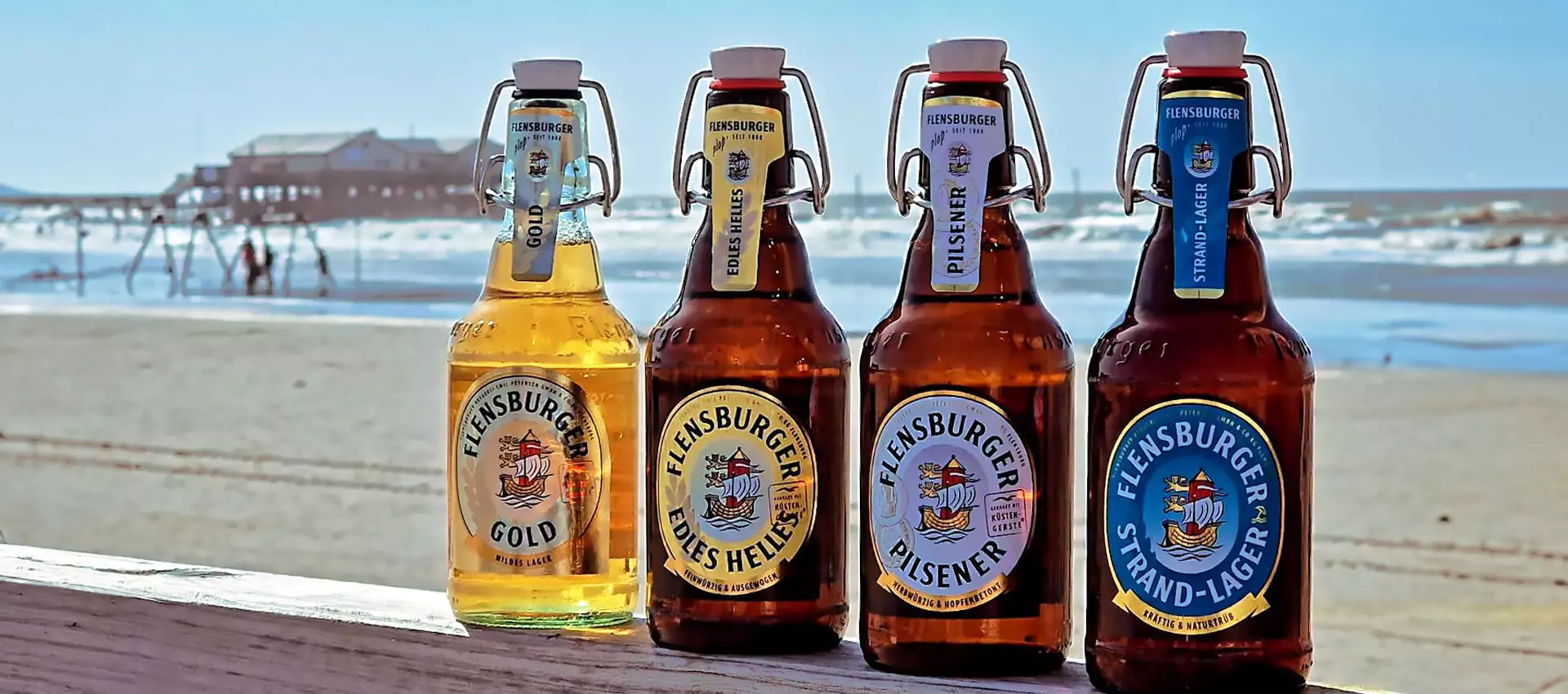 Vier FLENS Flaschen aufgereiht nebeneinander auf einer Brüstung vorm Strand.