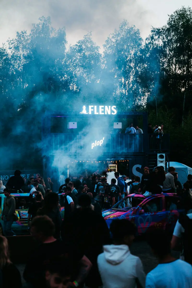 Butcher-Jam-Festival 2026 bei Dämmerung mit Menschenmenge, blauem Nebel, DJ-Bude "FLENS"-Leuchtschild und bunt bemalten Autos.