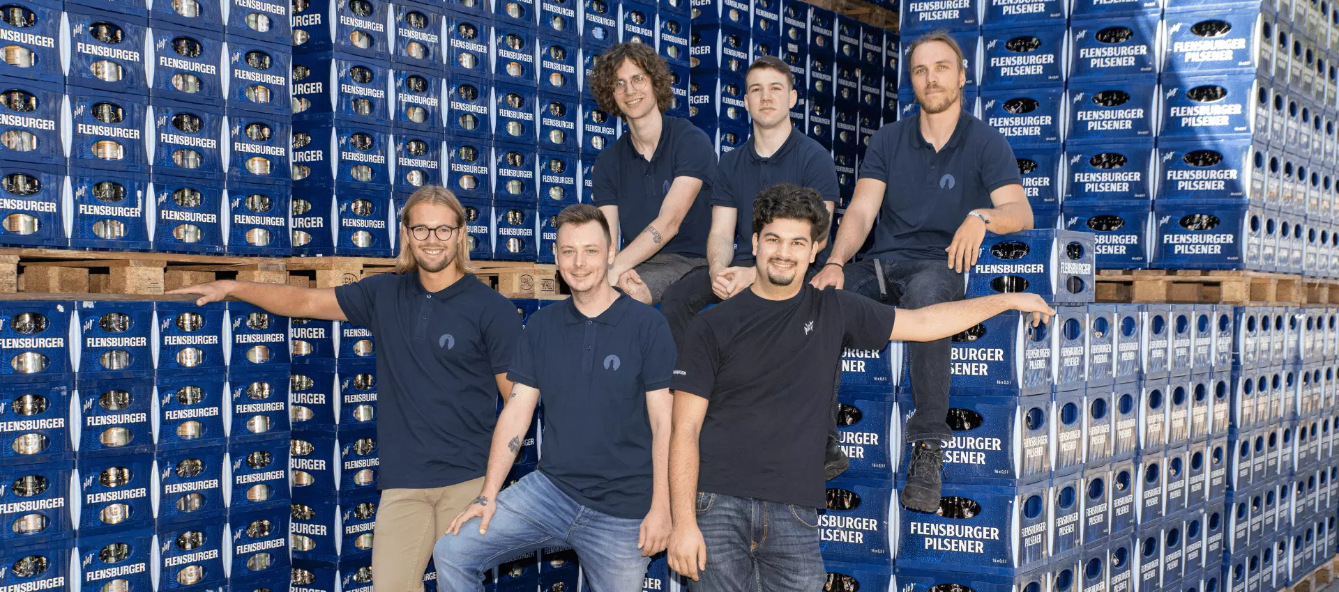 Fünf Flensburger Mitarbeiter, in blauen FLENS-Shirts vor Flensburger Kisten.