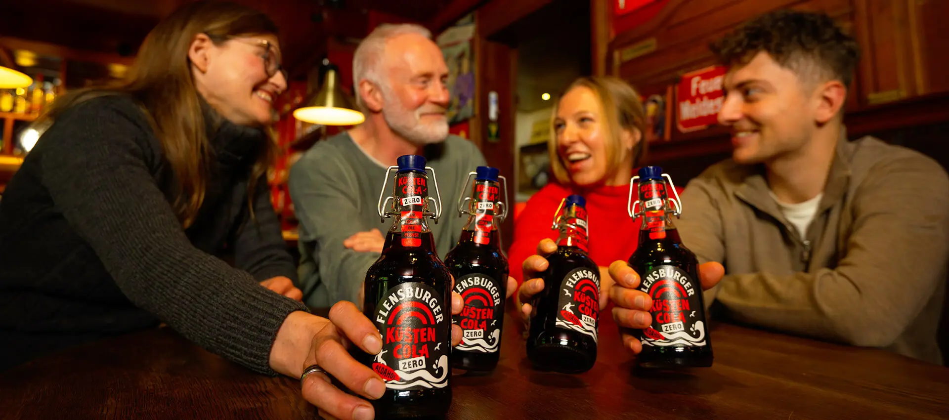 Vier Freunde in Kneipe stoßen mit Flensburger Küstencola-Zero-Flaschen an, gemütliche Bar-Atmosphäre.