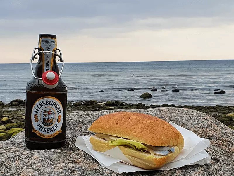 FLENS Pils mit einem Fischbrötchen, am Strand.