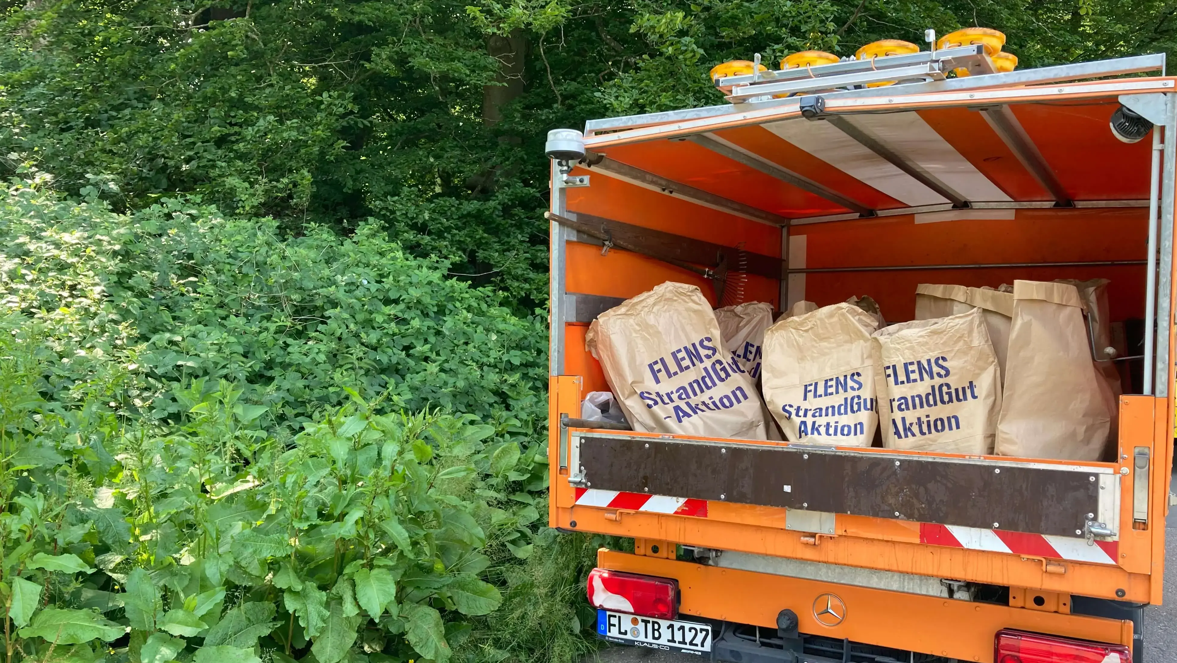 Oranger LKW mit geöffneter Ladefläche voller Säcke von der FLENS Strandreinigungsaktion in grüner Umgebung.
