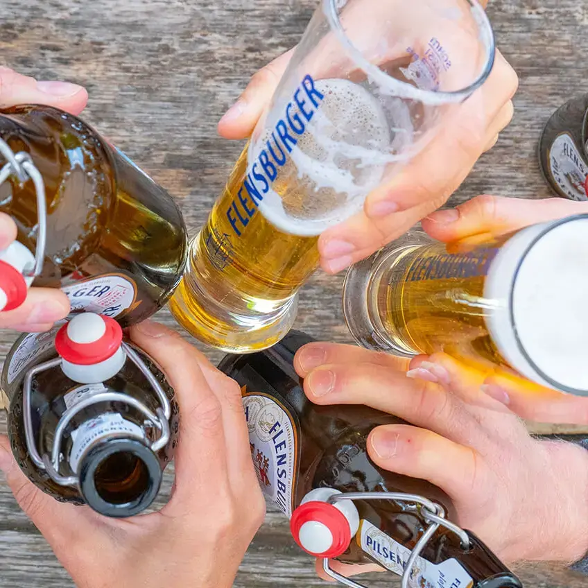 Freunde stoßen mit Flensburger Biergläsern und Bügelflaschen auf hölzernem Tisch an.