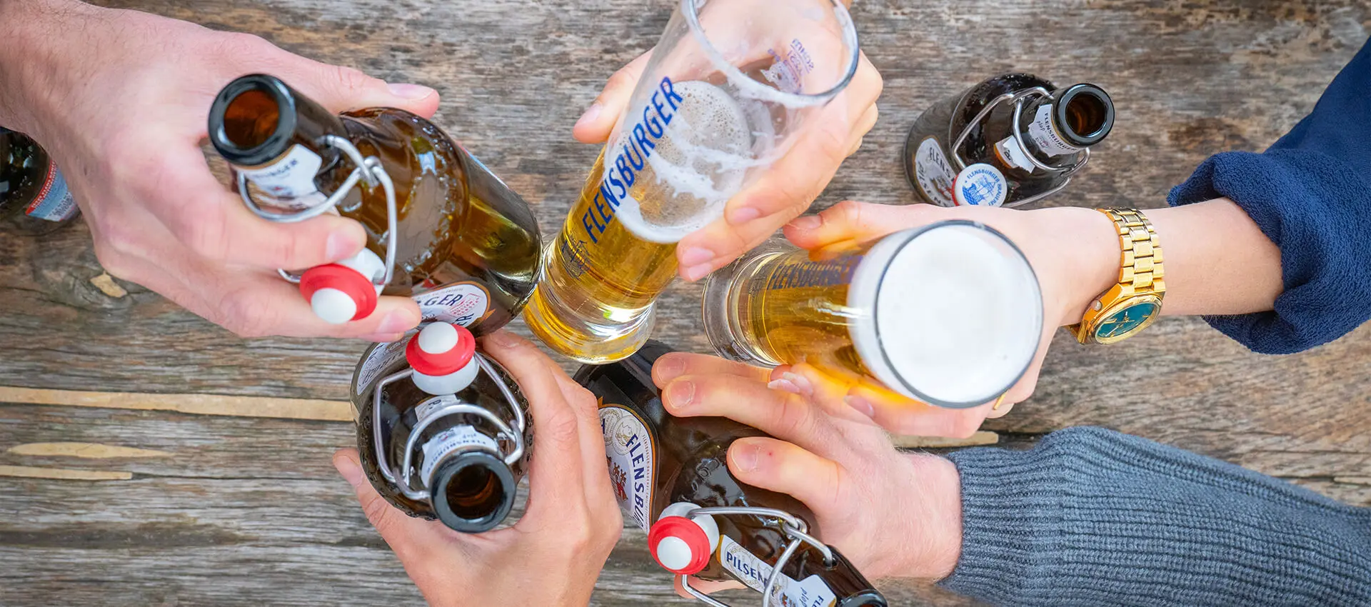 Freunde stoßen mit Flensburger Biergläsern und Bügelflaschen auf hölzernem Tisch an.