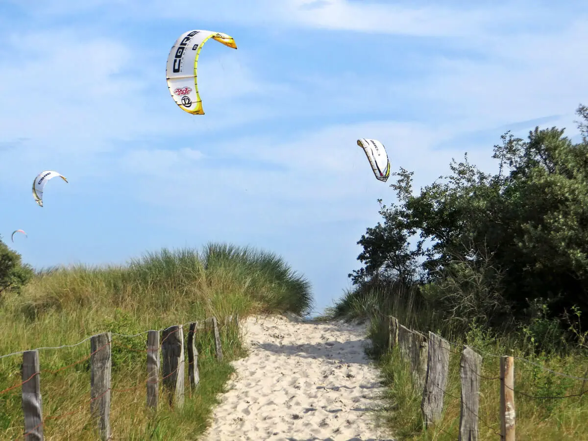 Sandweg durch Dünen zum Strand von Pelzerhaken mit Kitesurfern am blauen Himmel und Holzzaun.