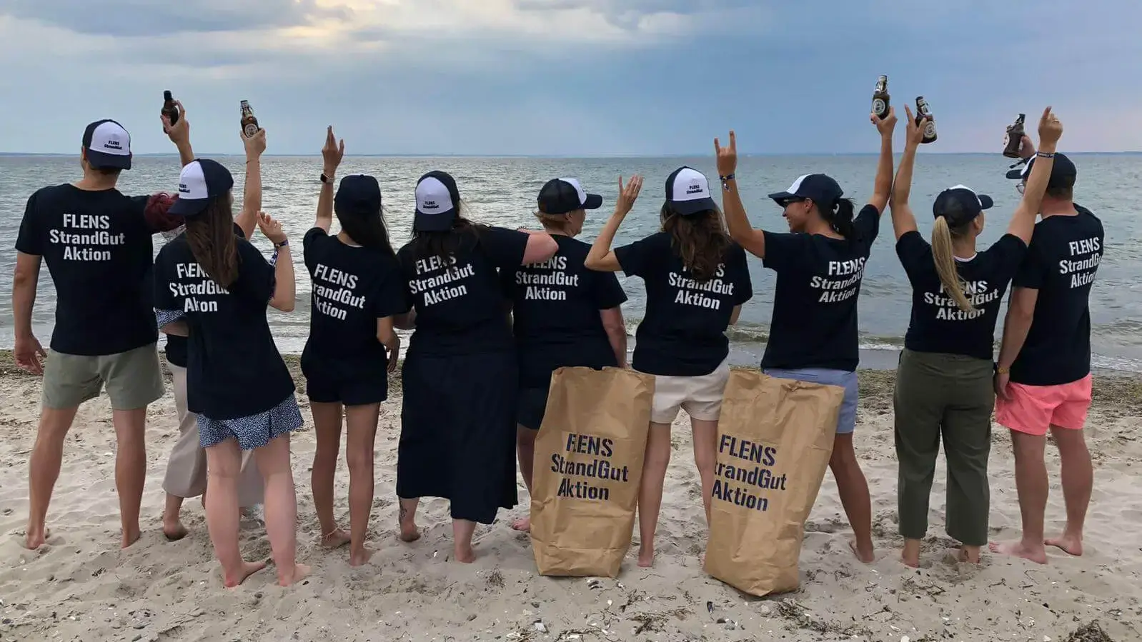 Gruppe von Menschen in StrandGut-Shirts mit FLENS Flaschen am Meer bei einer Strandreinigungsaktion.