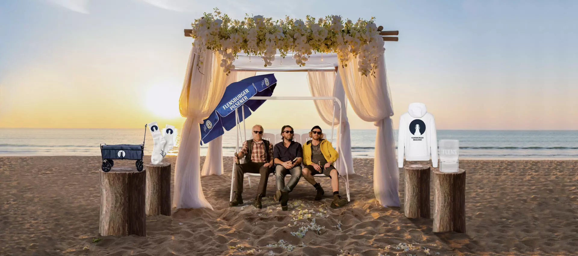 Bildmontage mit weißem Baldachin am Strand, darunter die drei Männer auf der Schaukel. Um sie herum sind einige Flensburger Merch-Artikel auf Pollern platziert.