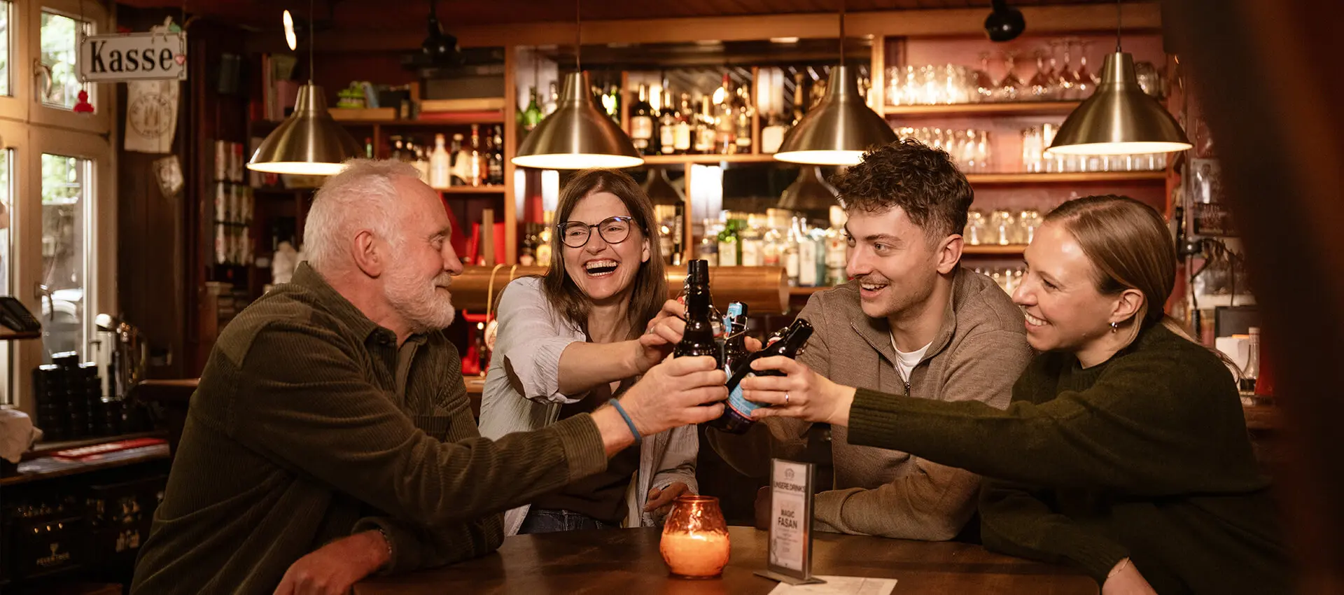 Vier Freunde stoßen mit FLENS Bierflaschen in gemütlicher Bar an, warmes Licht und Barregal im Hintergrund.