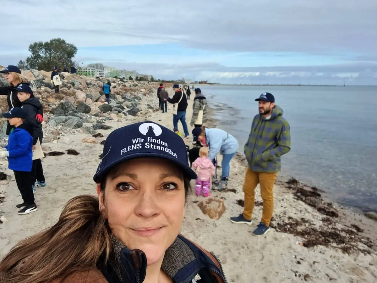 Frau mit Kappe macht Selfie am belebten Strand mit anderen Freiwilligen bei einer FLENS StrandGut Aktion.
