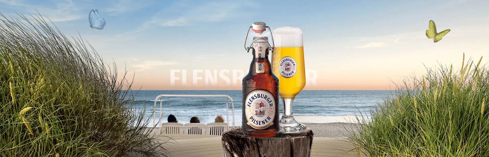 Spring-like beach scenery with wooden pole in the foreground, carrying a bottle and a glass of Flensburger Pilsener beer.