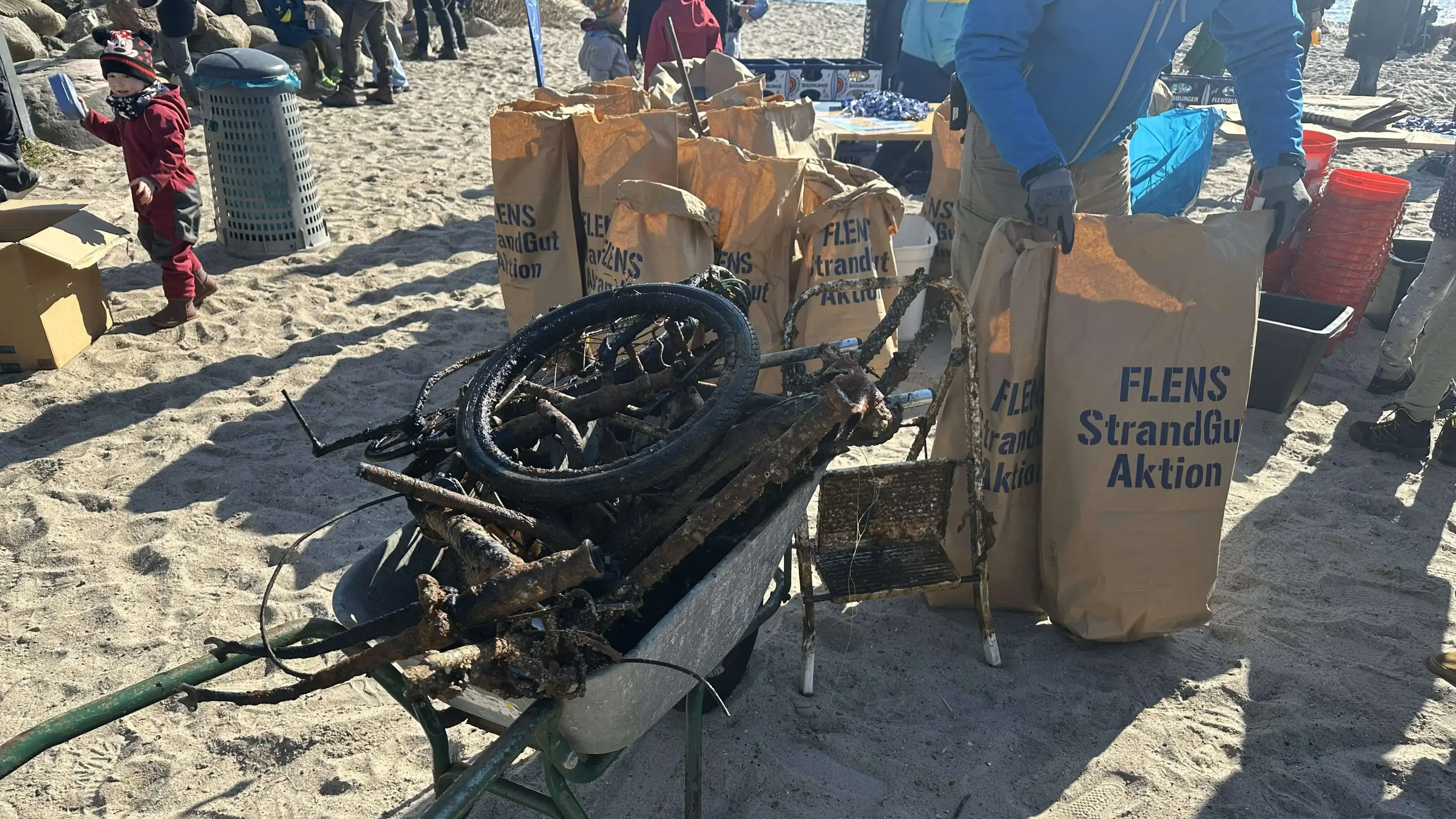Freiwillige sammeln Müll am Strand bei der FLENS StrandGut Aktion, rostiger gesammelter Müll im Vordergrund.