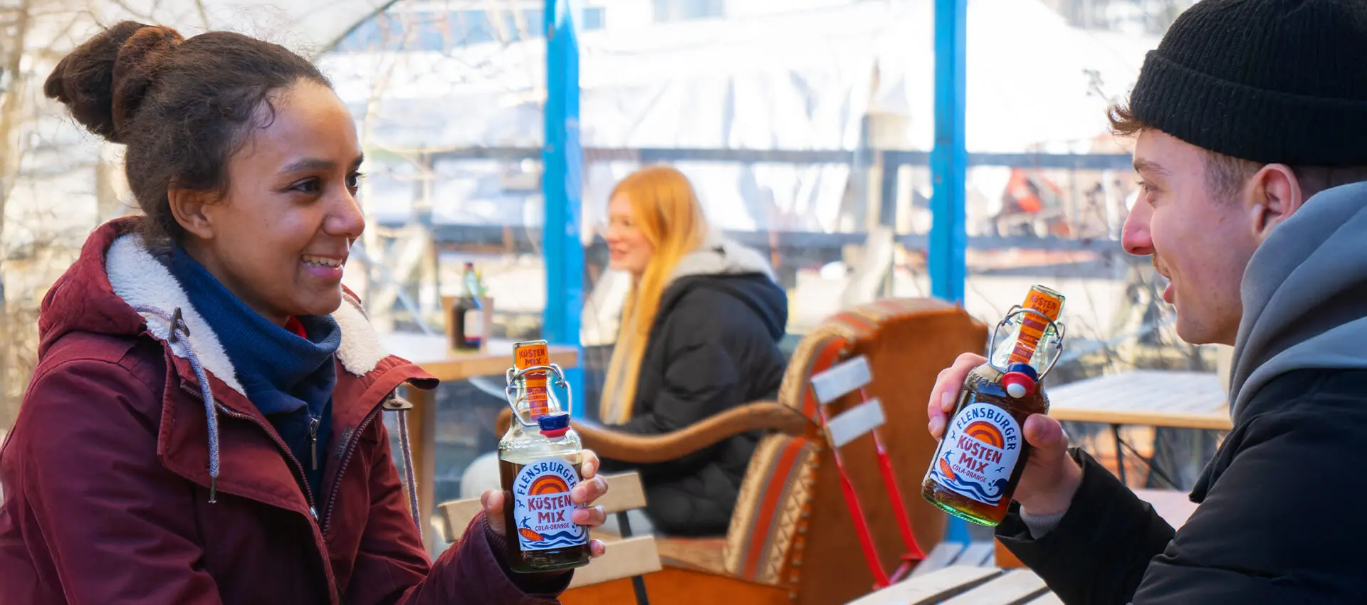Zwei Personen stoßen mit Flensburger Küstenmix-Flaschen an im winterlichen Außencafé.