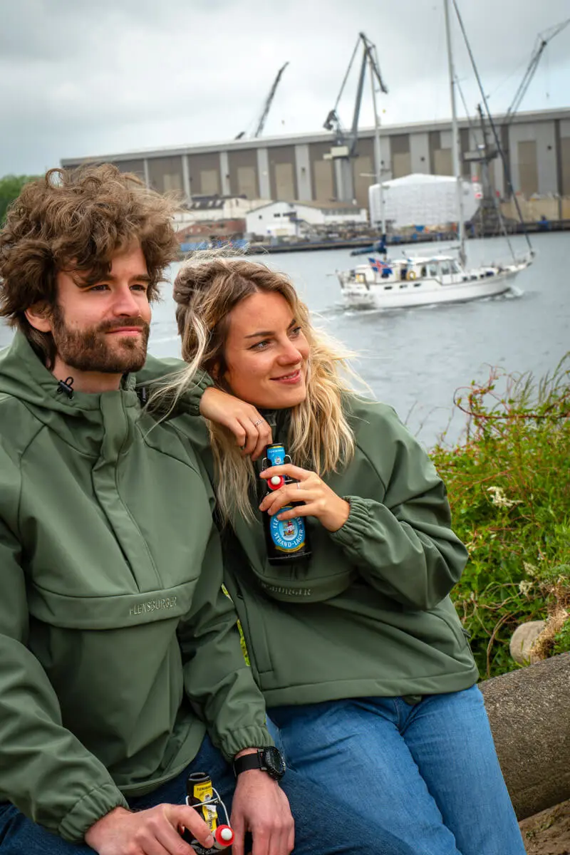 Pärchen in grünen Windbreaker-Jacken sitzt am Hafen, trinkt FLENS Bier, mit Segelboot und Werftkränen im Hintergrund.