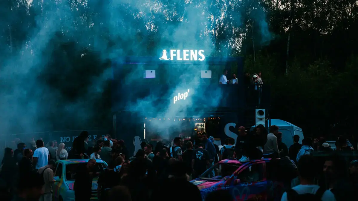 Butcher-Jam-Festival 2026 abends mit Rauch, Menschenmenge, beleuchteter 'FLENS' Bar und bunten, bemalten Autos.