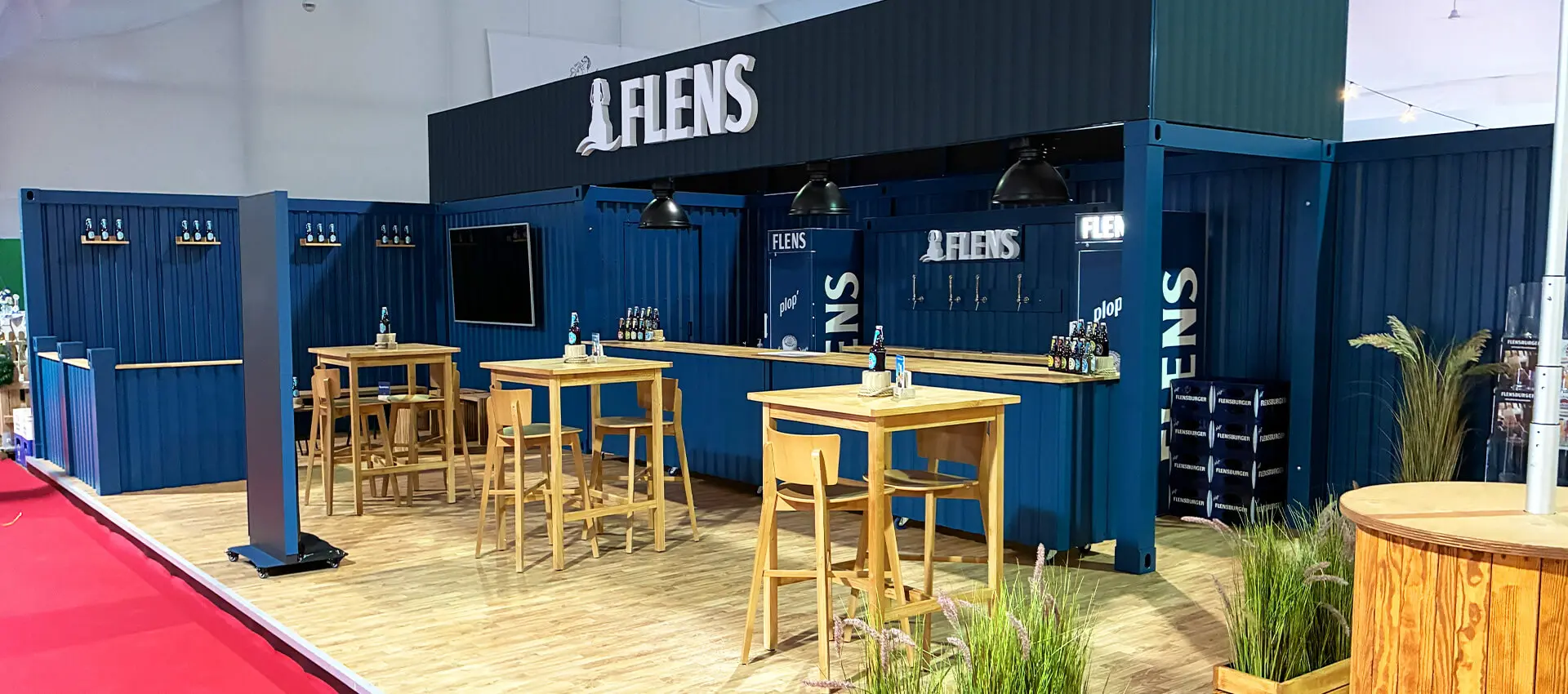 FLENS Messestand im blauen Container-Look mit Holztischen, Barhockern und Flensburger Bierflaschen.