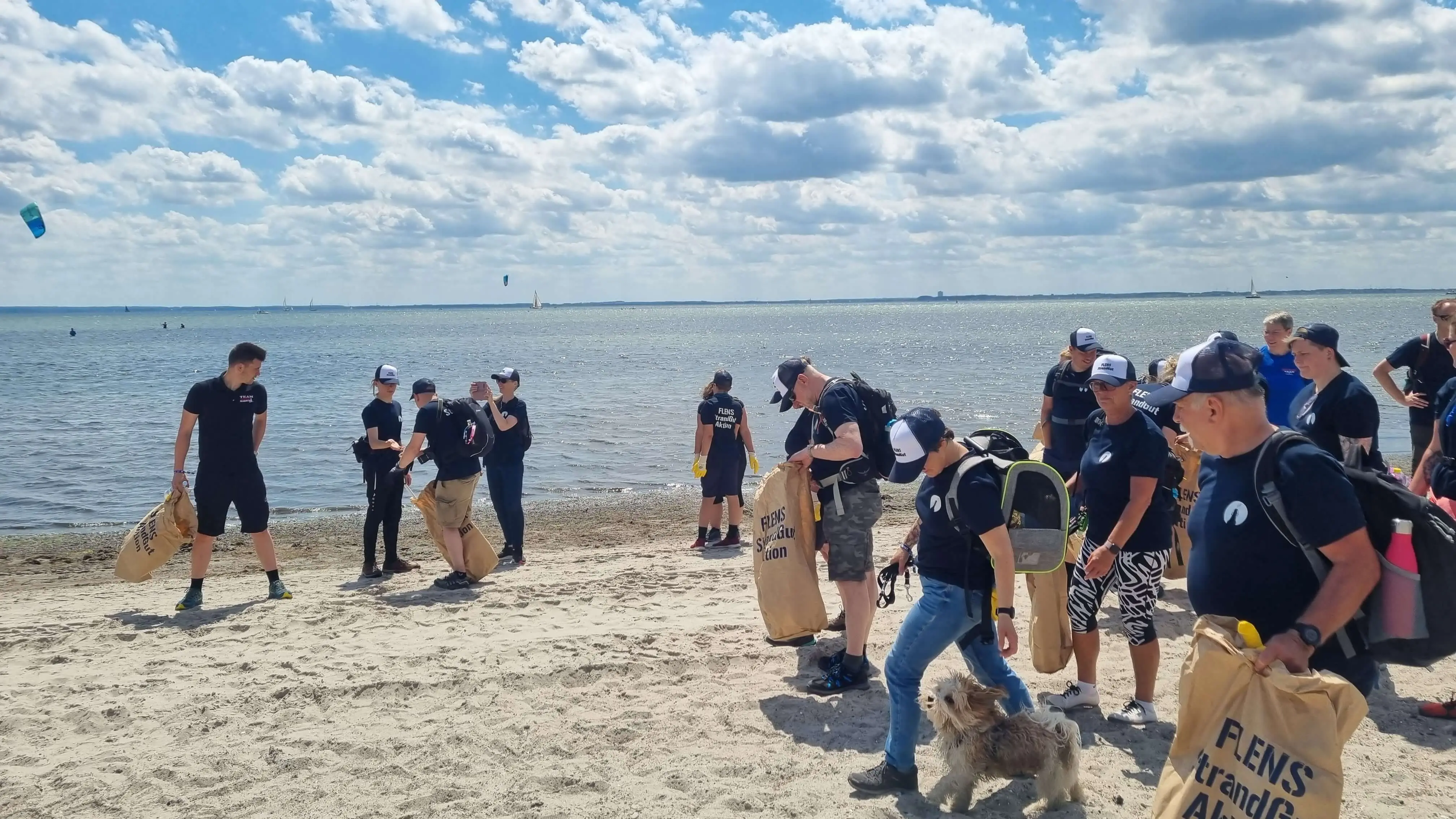 Gruppe von Menschen bei einer FLENS Strandreinigungsaktion mit Hund an einem sonnigen Tag.