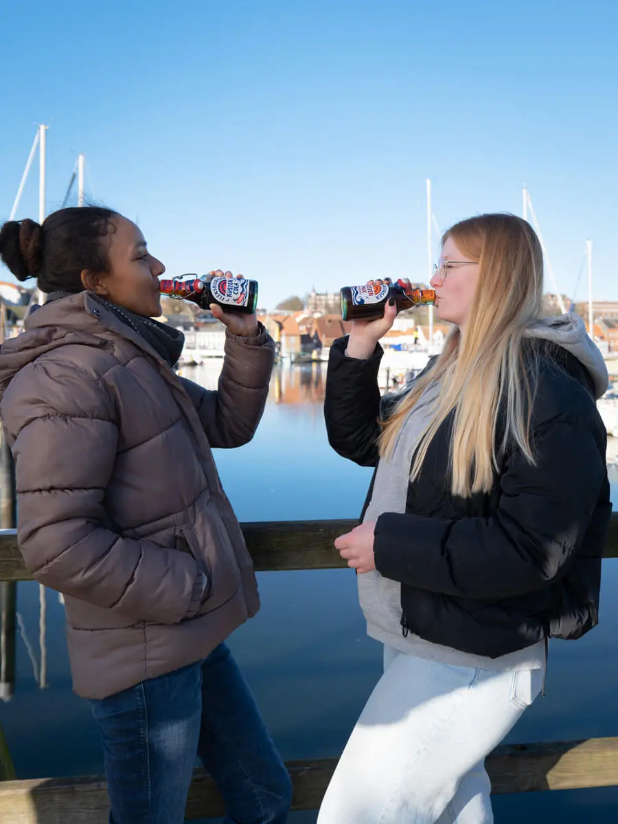 Zwei Frauen trinken Küstencola und Küstenmix am Hafenpier von Flensburg, Segelboote und Masten vor klarem blauem Himmel.