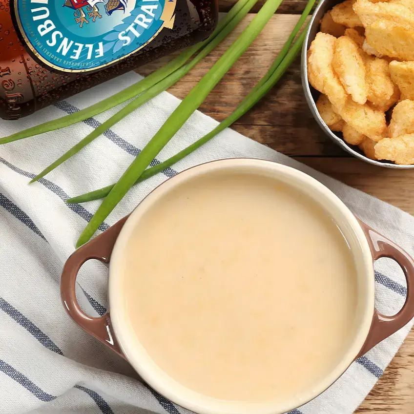 Flensburger Bierflasche neben cremiger Biersuppe, Schnittlauch, knusprigen Croutons und Löffel auf Holztisch.