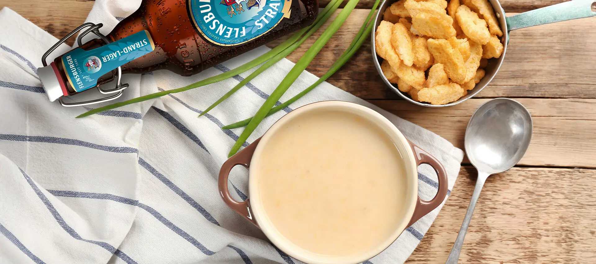 Flensburger Bierflasche neben cremiger Biersuppe, Schnittlauch, knusprigen Croutons und Löffel auf Holztisch.
