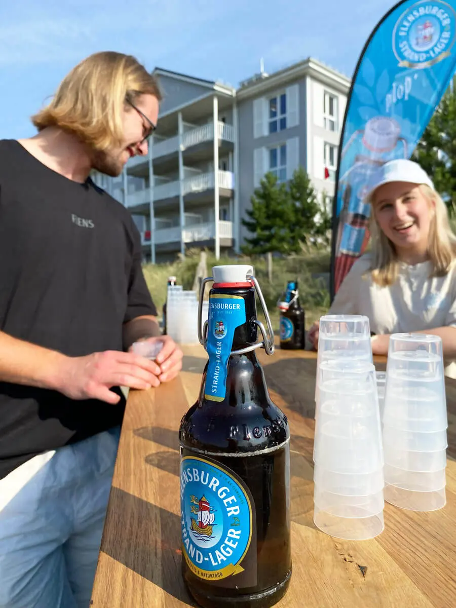 Flensburger Strand-Lager Bierflasche auf Holztheke mit Stapel Plastikbecher, Strand und Personen im Hintergrund.
