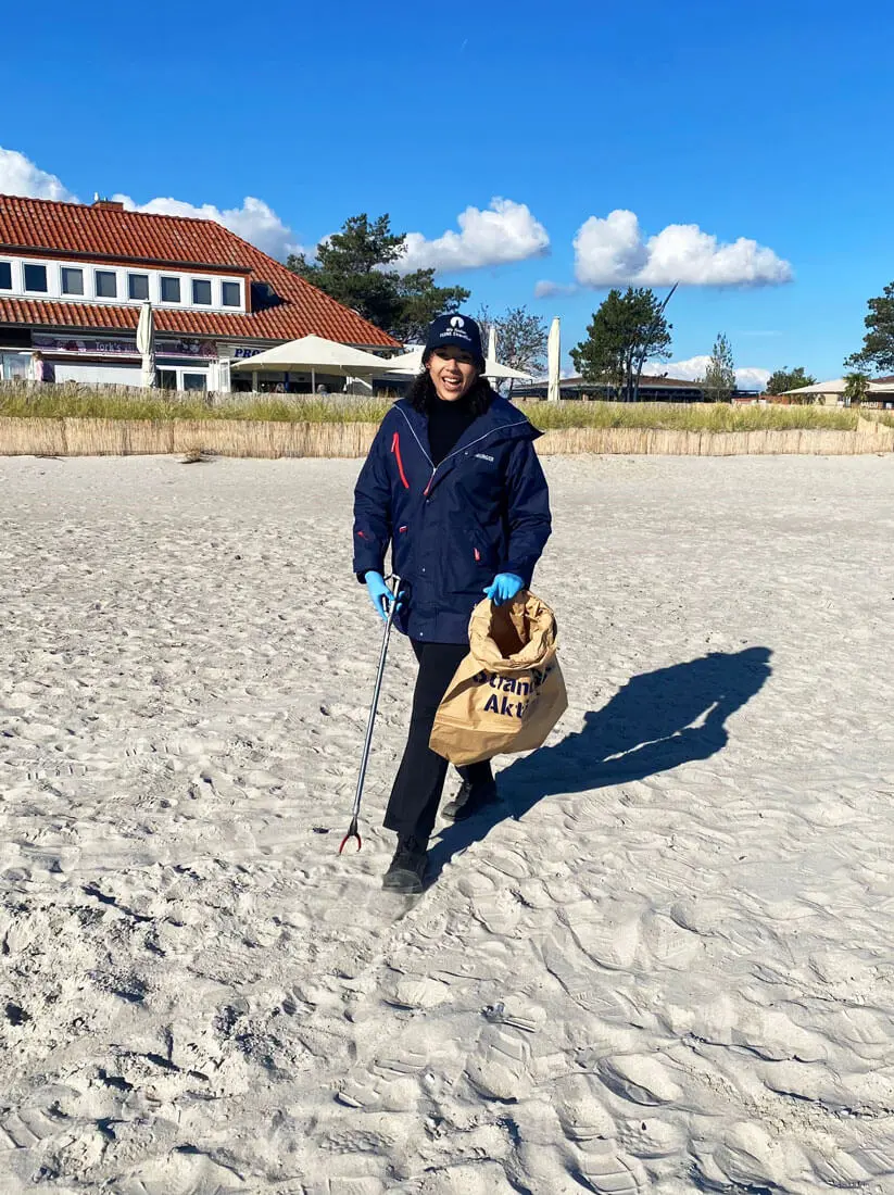 Frau sammelt Müll am Strand mit Greifer und Müllbeutel im Rahmen einer FLENS Strandreinigungsaktion bei sonnigem Wetter.