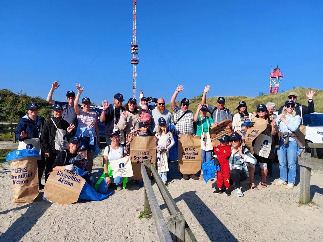 Gruppe von Freiwilligen mit Säcken bei FLENS Strandreinigungsaktion unter blauem Himmel.