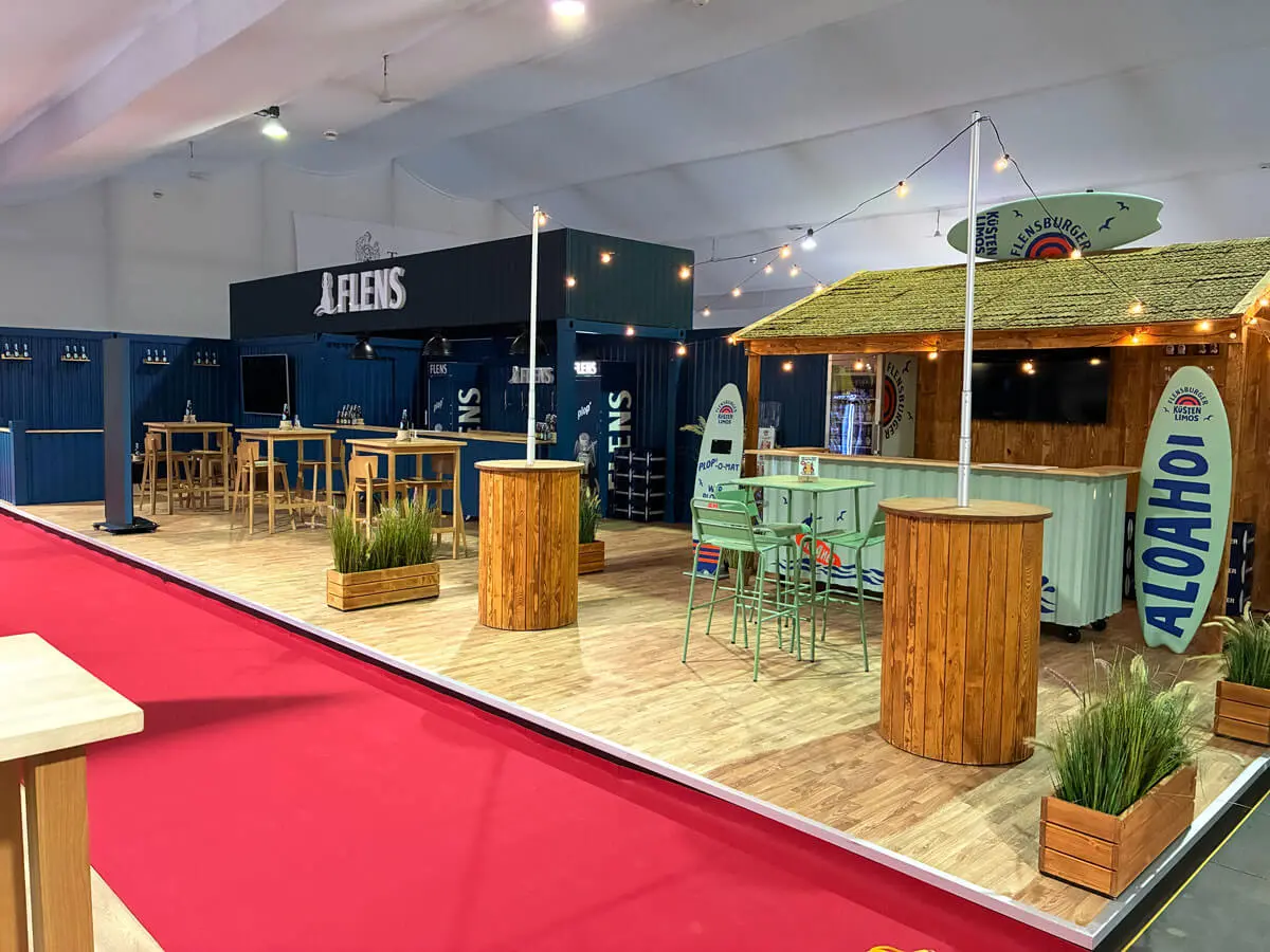 Indoor-Messestand mit Holztischen, Stehbar, Lichterketten, Küstenpflanzen, blauer Containerwand und Aloahoi-Deko.