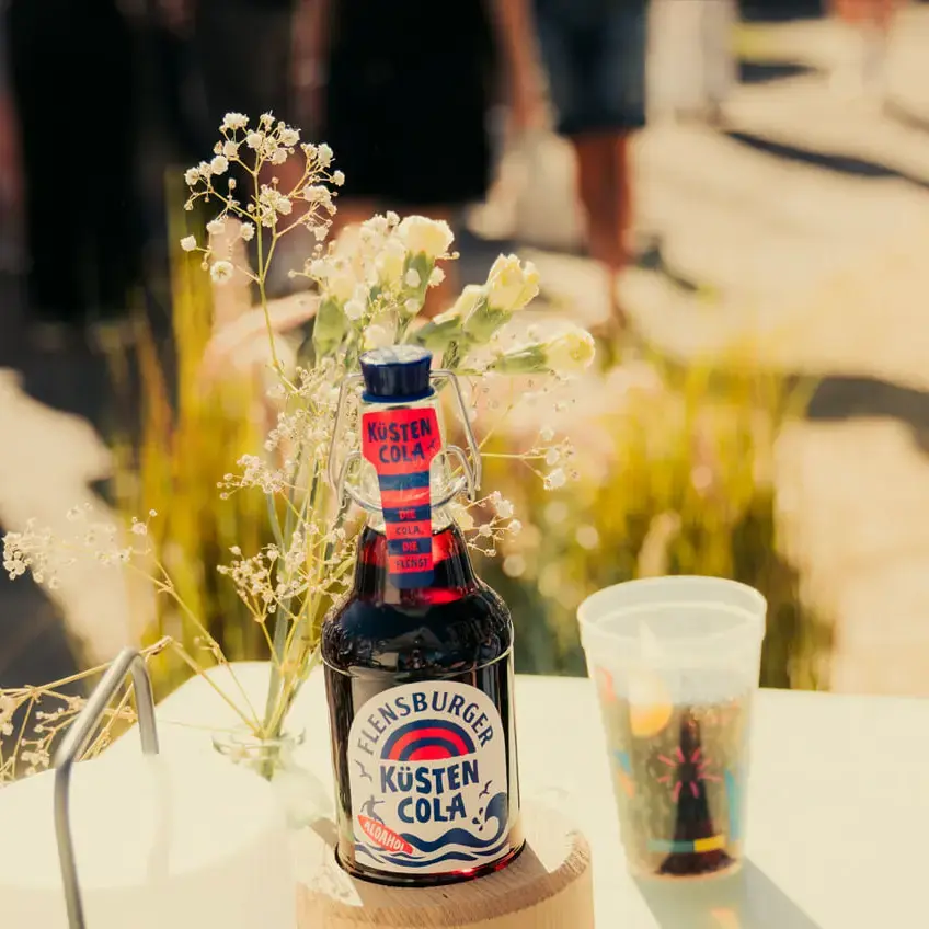 Flensburger Küstencola-Flasche auf Tisch mit Plastikbecher und Blumen auf der Kieler Woche 2026.