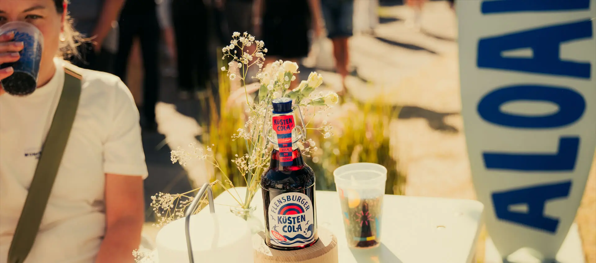 Flensburger Küstencola-Flasche auf Tisch mit Plastikbecher und Blumen auf der Kieler Woche 2026.