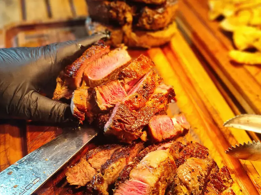 Person schneidet medium gegrilltes Ribeye-Steak auf einem Holzbrett, saftige Fleischstücke und Messer sichtbar.