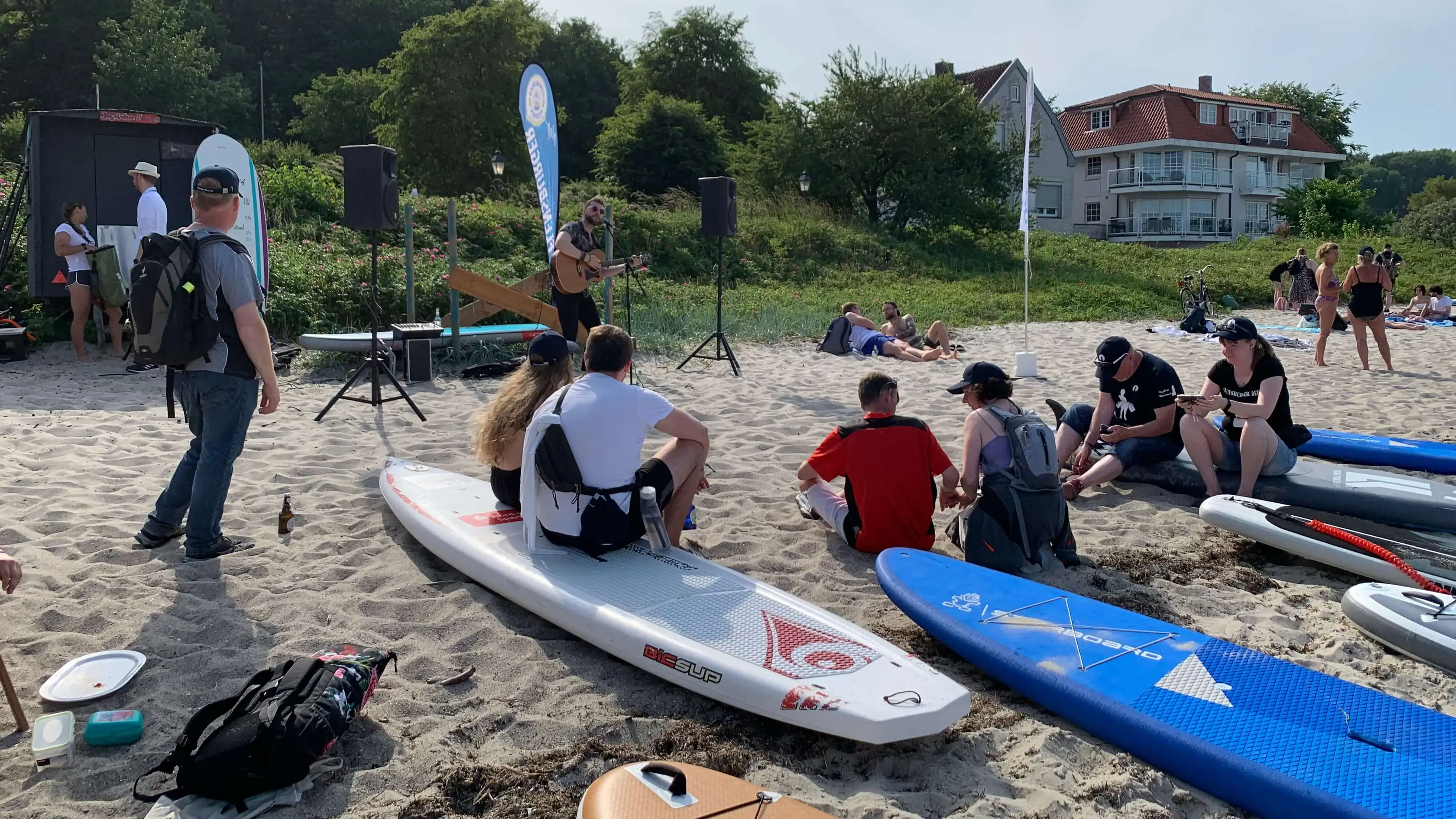 Live-Musik und entspannte Zuschauerinnen und Zuschauer am Strand mit Stand-Up-Paddleboards nach getaner Arbeit bei der FLENS StrandGut Aktion.
