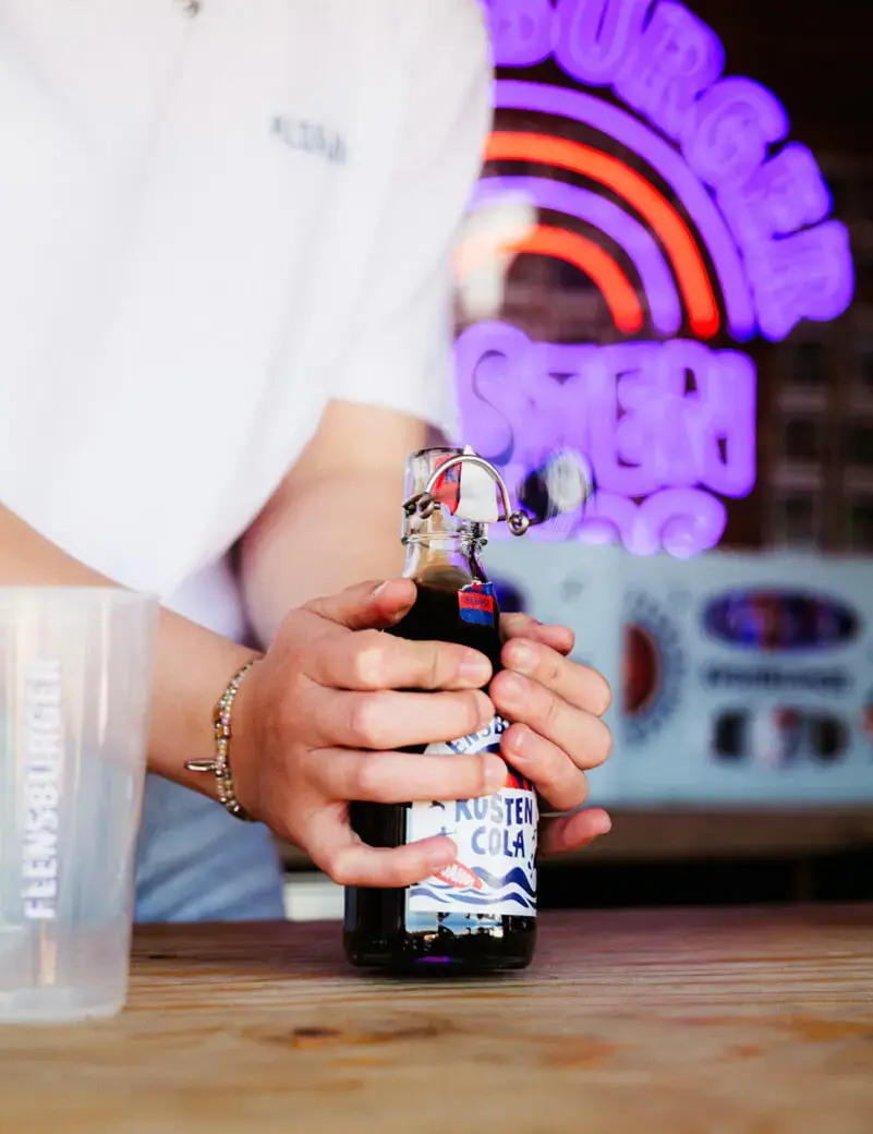 Eine Person ploppt eine Flasche Flensburger Küstencola, im Hintergrund das Flensburger-Küstenlimo-Leuchtzeichen.