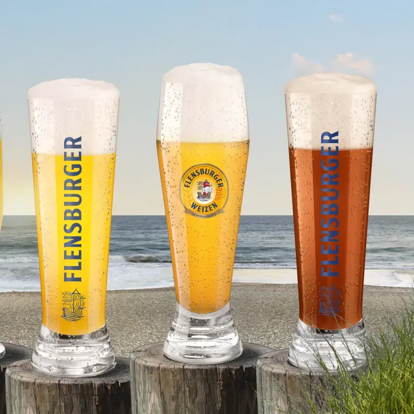 Vier Gläser mit unterschiedlich hellen Bieren auf Pollern am Strand.
