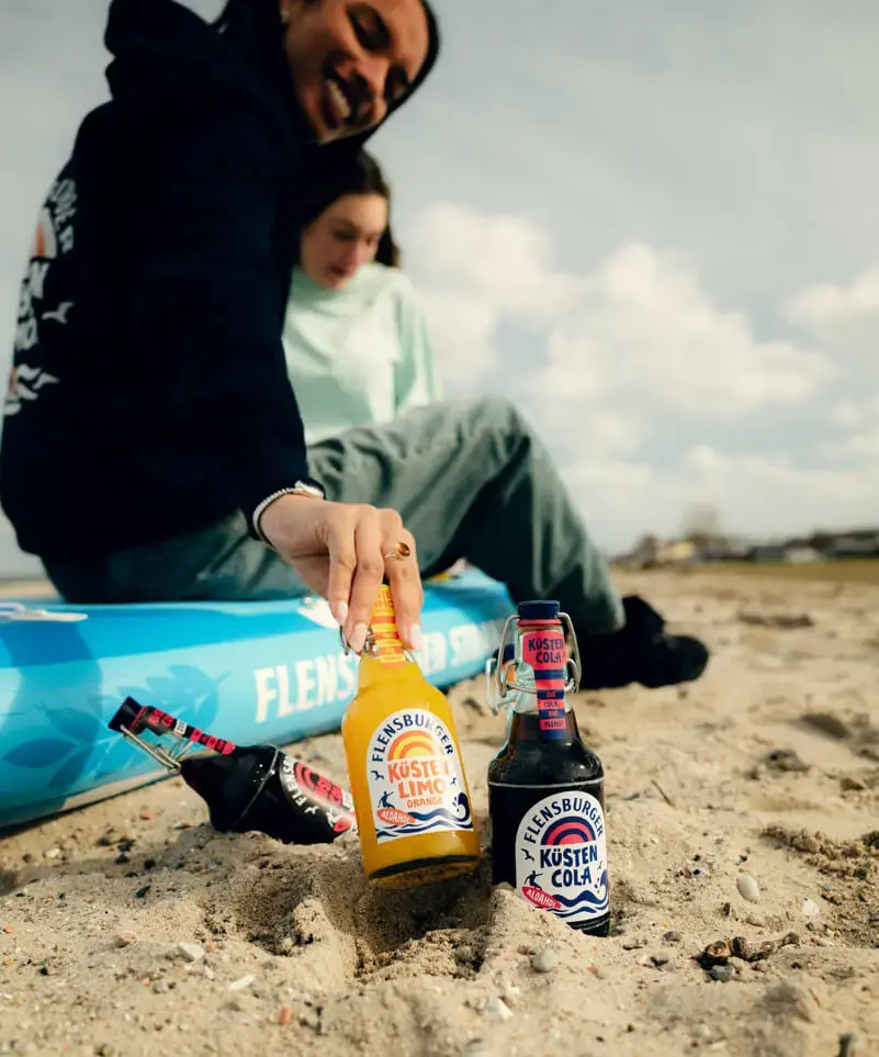 Eine junge Frau greift nach einer Flensburger Küstenlimo- und Küsten-Cola-Flasche im Sand am Strand neben einem SUP-Board.