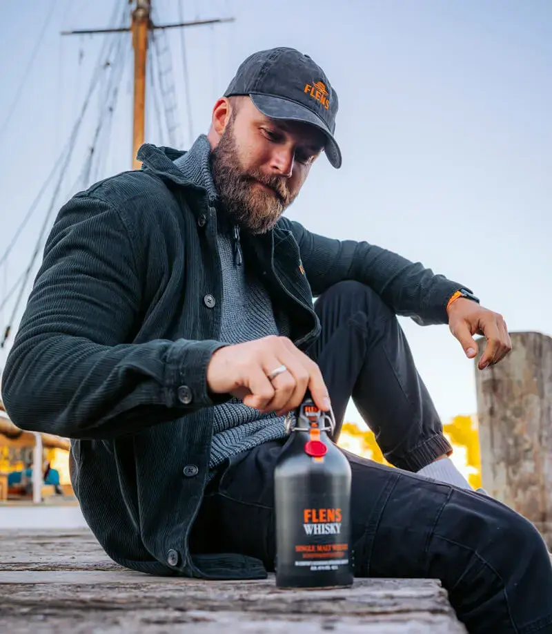 Mann im Whisky-Merch sitzt am Hafen mit der FLENS-Whisky-Flasche, der Schifffahrtsedition aus 2025.